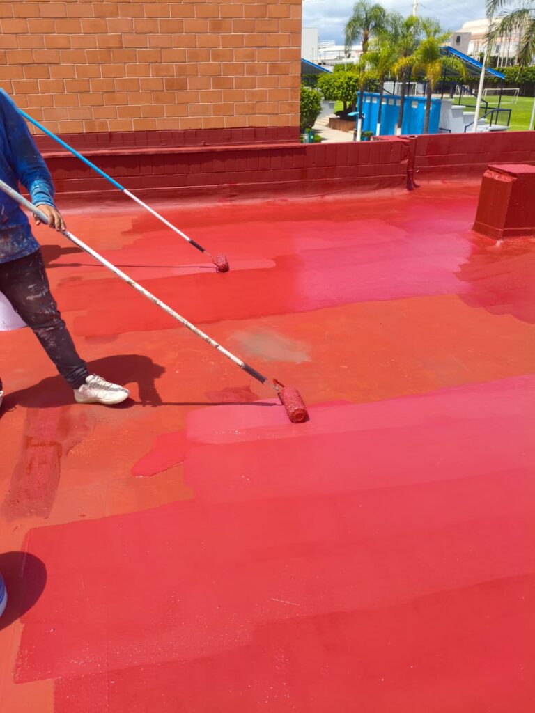 Una persona pinta el techo de rojo con rodillo cerca de un muro de ladrillo. Hay árboles y un edificio al fondo.