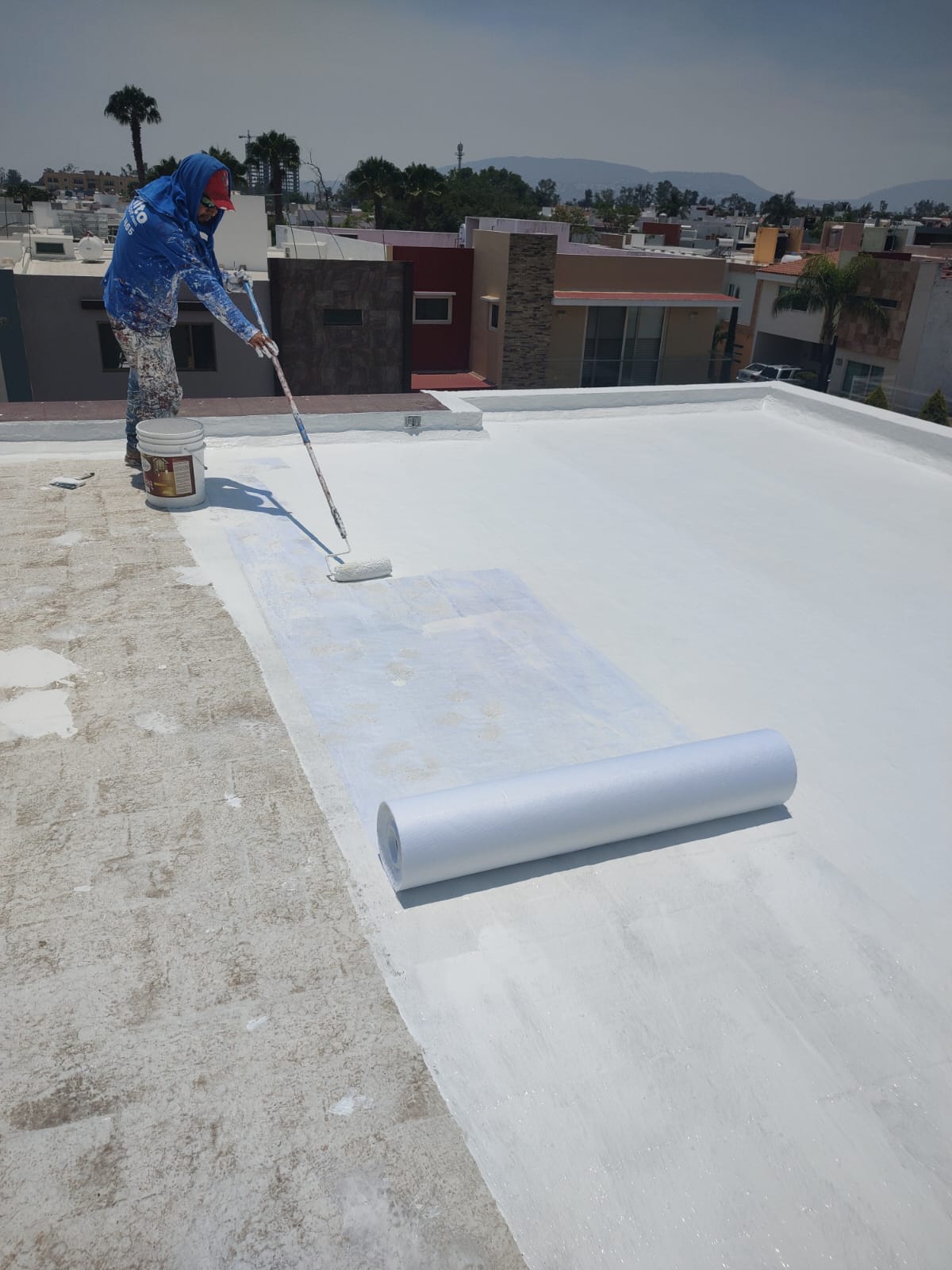 Un persona pinta un techo blanco en una azotea. Herramientas cercanas. Casas y palmeras en el fondo sin edificios reconocibles.